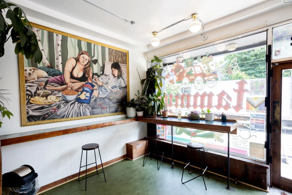 Bright and cozy café interior with large front windows, potted plants on a wooden counter, a bar stool seating area, and a bold wall painting depicting two women lounging with snacks and a dog, creating a quirky and inviting vibe.